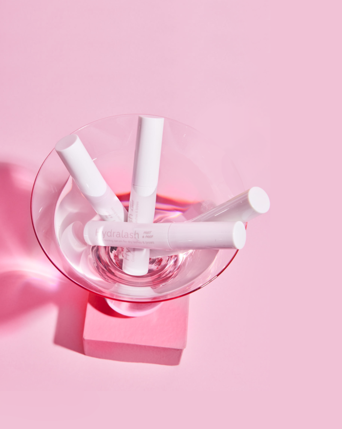 cocktail glass with clear liquid and 4 tubes of hydralash immersed in it on a pink background