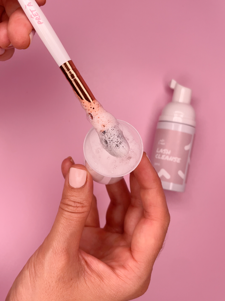 hand holding cup with bubbling cleanser being mixed by a small brush with a pink and white bottle in the background on a pink backdrop